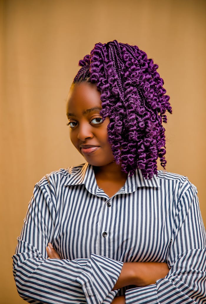 Portrait of a confident woman with vibrant purple braids and striped shirt against a neutral background.
